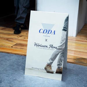 Puede incluir: Un letrero publicitario blanco con el texto "CODA Coffee x H&eacute;risson Flora Flower/Wedding/Party" en azul y negro. El letrero tambi&eacute;n incluye el texto "Morphing To Flower Blossom". El letrero est&aacute; sobre un suelo gris.
