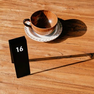 May include: A wooden teacup with a dark brown grain pattern sits on a white scalloped saucer. Next to the saucer is a black rectangular table number sign featuring the white number 16 printed on the front face of the upright display.