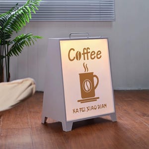 Può includere: Cartello da caffetteria a forma di A con display illuminato. Il cartello è grigio con una maniglia in alto. Il display mostra la parola "Coffee" e una grafica di tazza di caffè marrone. Il testo "KA FEI XIAO DIAN" è sotto.