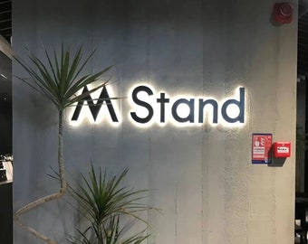 Pode incluir: Uma placa preta com luzes brancas ao redor das bordas lê "M Stand" em uma parede de concreto cinza. Uma planta verde está em primeiro plano.
