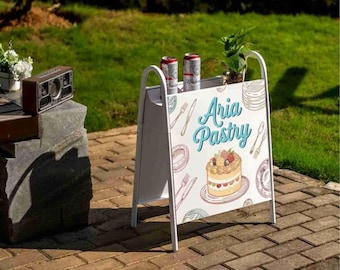 Könnte beinhalten: A-förmiges Werbeschild für "Aria Pastry" mit weißem Rahmen und einem Schild mit einer Kuchenillustration und Besteck. Zwei Dosen und eine kleine Pflanze stehen oben auf dem Schild. Das Schild steht auf einer Ziegeloberfläche.