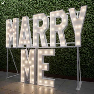 Op de afbeelding: Grote witte marquee letters met de tekst "MARRY ME" met verlichte lampen. De letters zijn gemonteerd op een wit metalen frame, tegen een groene heg achtergrond. De letters zijn verlicht, waardoor een romantische sfeer ontstaat.