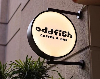 Puede incluir: Un letrero redondo e iluminado para "oddfish COFFEE & BAR" está montado en una pared texturizada. El letrero tiene un marco y un soporte negros, con el texto en negrita negra. Se ven plantas verdes en la parte inferior de la imagen.