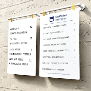 May include: Two white rectangular menu boards suspended from white horizontal bars with yellow clips. The boards feature black text listing food items like Margherita, Calzone, and Spicy Nduja with prices, and the Bru Coffee Roasters logo at the top.