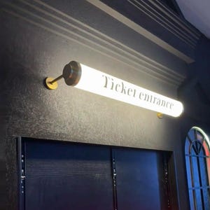 May include: A rectangular, illuminated sign with the words "Ticket entrance" in black lettering. The sign is mounted on a dark wall with gold-colored hardware. The sign is a bright white color.