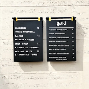 May include: Two rectangular black menu boards with white text mounted on a wall. The left board lists Margherita, Calzone, Spicy Nduja, and Hazelnut Pesto. The right board features the text The Good Life and lists various pizza options.