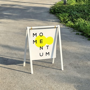 May include: A white A-frame sidewalk sign with a rectangular panel suspended from a top bar. The panel features the black text MOMENTUM arranged in a grid, with two overlapping bright yellow circles positioned behind the letters O and M.