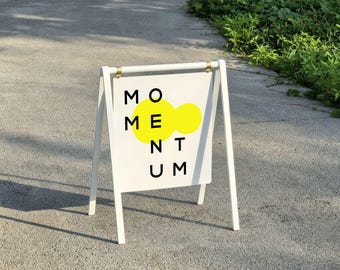 May include: A white A-frame sidewalk sign with a rectangular panel suspended from a top bar. The panel features the black text MOMENTUM arranged in a grid, with two overlapping bright yellow circles positioned behind the letters O and M.