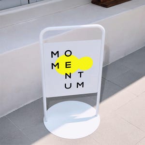 Op de afbeelding: Een wit metalen A-bord voor op de stoep met een ronde voet. Het rechthoekige bordoppervlak bevat de zwarte tekst MOMENTUM in een raster, met een felgele organische vorm achter de letters O, M, E, N, T en U geplaatst.