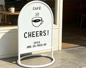 Puede incluir: Un letrero de café blanco arqueado con texto negro y un gráfico de taza de café. El letrero dice "CAFÉ", un icono de taza de café y "CHEERS!". Debajo, indica "OPEN AM8:30-PM20:00". El letrero está sobre un marco de metal blanco.