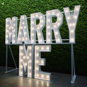 以下が含まれることがあります： 「MARRY ME」と書かれた大きな白い電光文字。文字は電球で照らされ、白い金属フレームに取り付けられています。背景は緑の生け垣です。プロポーズや結婚式のための装飾品。