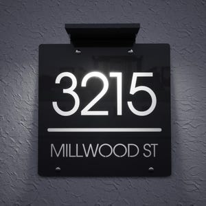 May include: A black illuminated address sign displaying the number "3215" in white, above the text "MILLWOOD ST". The sign is mounted on a textured grey wall and has a black top.