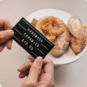 May include: A black rectangular sign with white horizontal lines and white text that reads HANDMADE CERAMICS $12.00. The sign is held in front of a white plate containing a golden-brown salted pretzel and two pieces of crusty bread.