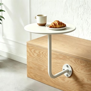 May include: A white, round side table with a curved, metal support attached to a wooden cabinet. A mug and a croissant on a small plate sit on the table. The table is approximately 12 inches in diameter.