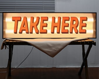 May include: A lit sign with the words "TAKE HERE" in orange lettering on a white background. The sign is resting on a wooden table.