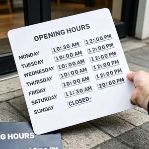 May include: A white square sign with black text displaying OPENING HOURS for each day of the week. The schedule lists Monday through Friday with 10:00 AM to 12:00 PM, Saturday with 11:30 AM to 20:00 PM, and Sunday marked as CLOSED- in a clean font.