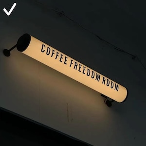 Peut inclure: Un enseigne cylindrique et lumineuse avec les mots "COFFEE FREEDOM ROOM" en lettres noires. L'enseigne est fixée sur un mur sombre avec des fixations noires. L'enseigne émet une lueur chaleureuse et accueillante.