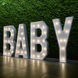 Może przedstawiać: Duże, podświetlane białe litery, które piszą "BABY" z ciepłymi białymi światłami. Litery są umieszczone na kafelkowej podłodze na tle zielonych liści. Światła są równomiernie rozmieszczone w każdej literze.