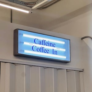 May include: A rectangular sign with the words "Caffeine Coffee In" in blue lettering. The sign is illuminated by three horizontal fluorescent lights. The sign is mounted on a white wall.