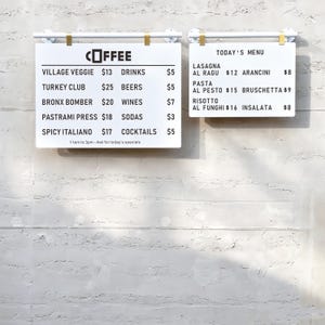 May include: Two white rectangular menu boards with black text hanging from white and gold metal bars. The left board lists coffee and food items like Village Veggie and Turkey Club with prices. The right board displays Today's Menu with Italian dishes.