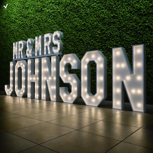 以下が含まれることがあります： 緑の生け垣の背景に、「MR & MRS JOHNSON」と書かれた大きな照明付きのマーキー文字。 温白色のライトが付いた白い文字は、お祝いの雰囲気を醸し出しています。 結婚式やイベントに最適です。
