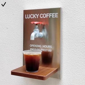 May include: A stainless steel wall-mounted coffee dispenser with a red-handled faucet. The sign reads "LUCKY COFFEE" and "OPENING HOURS PM13:00-PM17:00". A clear plastic cup of coffee sits on a small wooden shelf.
