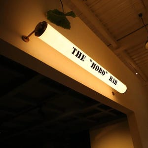 May include: A long, illuminated sign with the text "THE "HOBO" BAR" in black lettering. The sign is cylindrical and mounted on a wall. The sign is lit from within, casting a warm glow. A decorative leaf is visible above the sign.