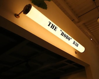 May include: A long, illuminated sign with the text "THE "HOBO" BAR" in black lettering. The sign is cylindrical and mounted on a wall. The sign is lit from within, casting a warm glow. A decorative leaf is visible above the sign.