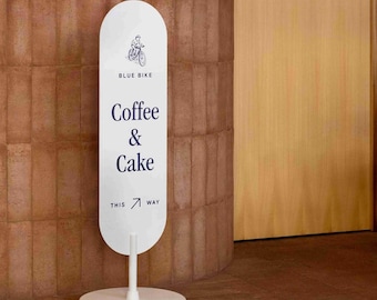 Puede incluir: Un letrero de suelo ovalado blanco con un gráfico azul oscuro de una persona en bicicleta, el texto "BLUE BIKE", "Coffee & Cake" y "THIS WAY" con una flecha. El letrero está montado sobre un poste vertical blanco y una base circular blanca.