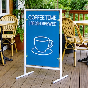 May include: A white metal A-frame sign with a blue panel. The panel features the text "COFFEE TIME | FRESH BREWED" and a white outline of a coffee cup and saucer. The sign is displayed outdoors near cafe tables.