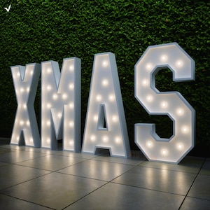 Könnte beinhalten: Große weiße Leuchtbuchstaben mit der Aufschrift "XMAS" mit beleuchteten Glühbirnen. Die Buchstaben stehen auf einer gefliesten Oberfläche vor einer grünen Hecke. Die Buchstaben sind beleuchtet und schaffen eine festliche Weihnachtsdekoration.