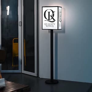 May include: A black and white sign post with the text "QUALITY KINGS" and a stylized "QK" logo. The sign is rectangular and illuminated, mounted on a tall, black pole. The sign is indoors, near a glass door.