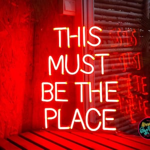 May include: Neon sign with the words "This Must Be The Place" in white letters on a red background.