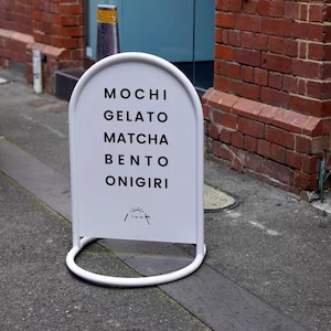 Op de afbeelding: Een wit, boogvormig sandwichbord op een stoep. Het bord vermeldt "MOCHI, GELATO, MATCHA, BENTO, ONIGIRI" in zwarte tekst. Het bord heeft een witte, afgeronde basis en staat voor een bakstenen muur.
