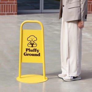 May include: A bright yellow A-frame sign with the words "Pluffy Ground" and a cartoon dog illustration. The sign is on a concrete surface.