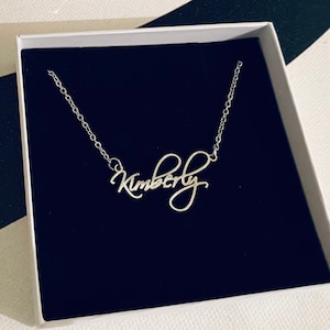 Peut inclure: Un collier en argent avec le nom "Kimberly" écrit en cursive. Le collier est dans une boîte blanche avec un revêtement en velours noir.