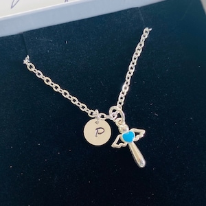 Puede incluir: Un collar de plata con un pequeño dije de ángel y un dije de la letra 'P'. El collar está en una caja de terciopelo negro.