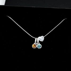 May include: A silver necklace with a delicate chain, featuring a small heart pendant and two round, colorful gemstone charms. One charm is amber-colored, the other is blue. The necklace is presented in a black gift box.