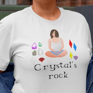 Può includere: Maglietta bianca con un'illustrazione di cartone animato di una donna seduta a gambe incrociate con cristalli intorno a lei. Il testo "Crystal's rock" è stampato sotto l'illustrazione.