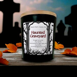 May include: A black candle with a wooden lid, labeled "Haunted Graveyard." The label features a decorative border with trees and a graveyard scene. The candle is 8 oz (227 g) and is a soy candle.