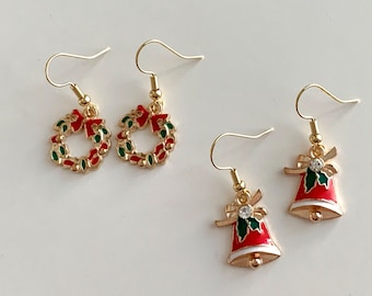 Op de afbeelding: Een paar gouden oorbellen met een kerstthema. Het ene paar heeft een rode en groene krans met een strik, en het andere paar heeft een rode bel met een groene strik en een witte kraal.