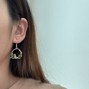 Puede incluir: Pendientes de aro dorados con un dise&ntilde;o de esmalte verde y naranja de dos p&aacute;jaros posados en una rama con hojas verdes.