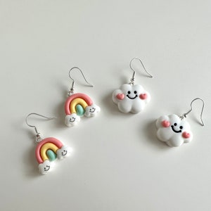 May include: A pair of rainbow and cloud earrings. The rainbow earrings are pink, yellow, and green with a white cloud on each end. The cloud earrings are white with a pink heart on each side and a smiley face in the center.