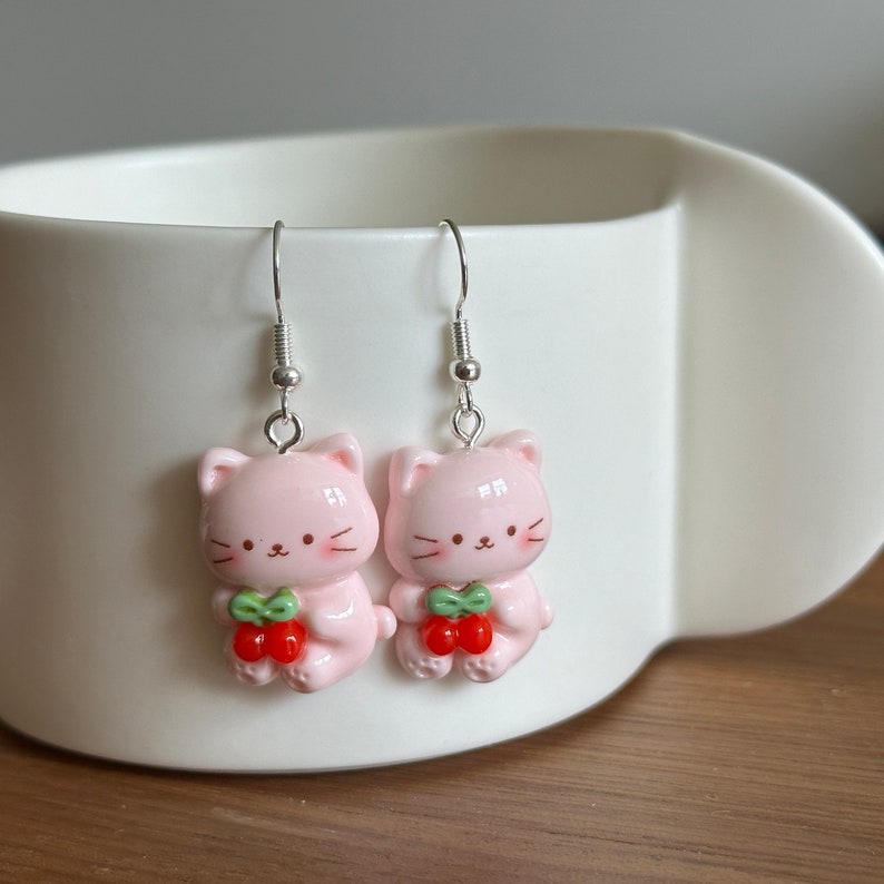 May include: A pair of pink cat-shaped earrings with silver hooks. Each cat holds a red cherry with green leaves. The earrings are a cute, whimsical accessory.
