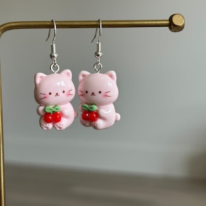 May include: A pair of pink cat-shaped earrings with silver hooks. Each cat holds a cluster of red cherries with green leaves. The earrings have a glossy finish and are designed in a cute, cartoon style.