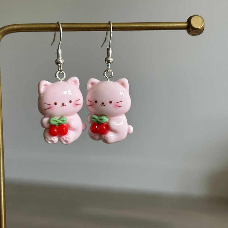 May include: A pair of pink cat-shaped earrings with silver hooks. Each cat holds a cluster of red cherries with green leaves. The earrings have a glossy finish and are designed in a cute, cartoon style.
