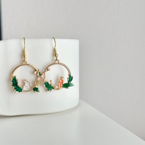 Puede incluir: Pendientes de aro dorados con un dise&ntilde;o de esmalte verde, rosa y blanco con un conejo y un p&aacute;jaro.
