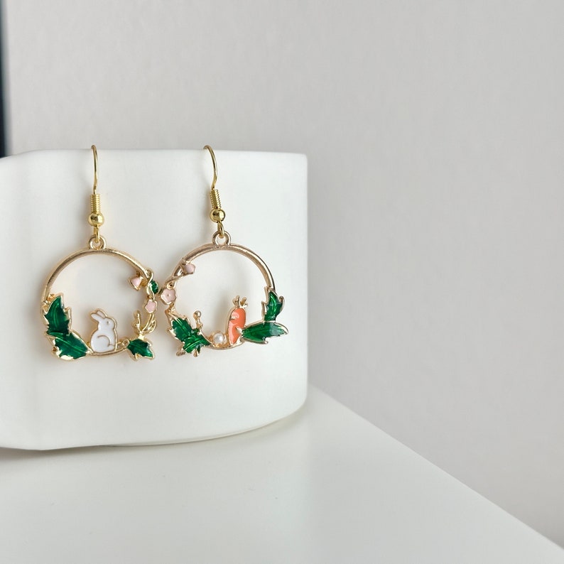 Puede incluir: Pendientes de aro dorados con un dise&ntilde;o de esmalte verde, rosa y blanco con un conejo y un p&aacute;jaro.