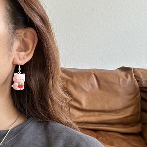 May include: Pink cat earrings with silver hooks. Each earring features a small, pink cat holding a red strawberry. The earrings are a cute and whimsical accessory.