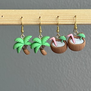May include: A set of four gold earrings featuring tropical themes. Two earrings are shaped like palm trees with green leaves and brown trunks. The other two earrings are shaped like coconuts with pink straws and green umbrellas.